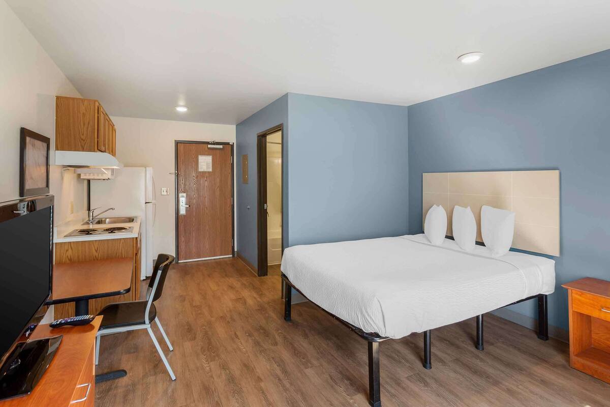 A hotel room with a bed, a desk with a chair, a TV, and a kitchenette.