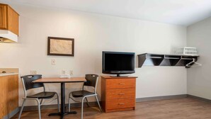 Een kleine, moderne kamer met een houten tafel en twee stoelen, een flatscreen televisie, een klein kastje en een wandplank met een mandje.