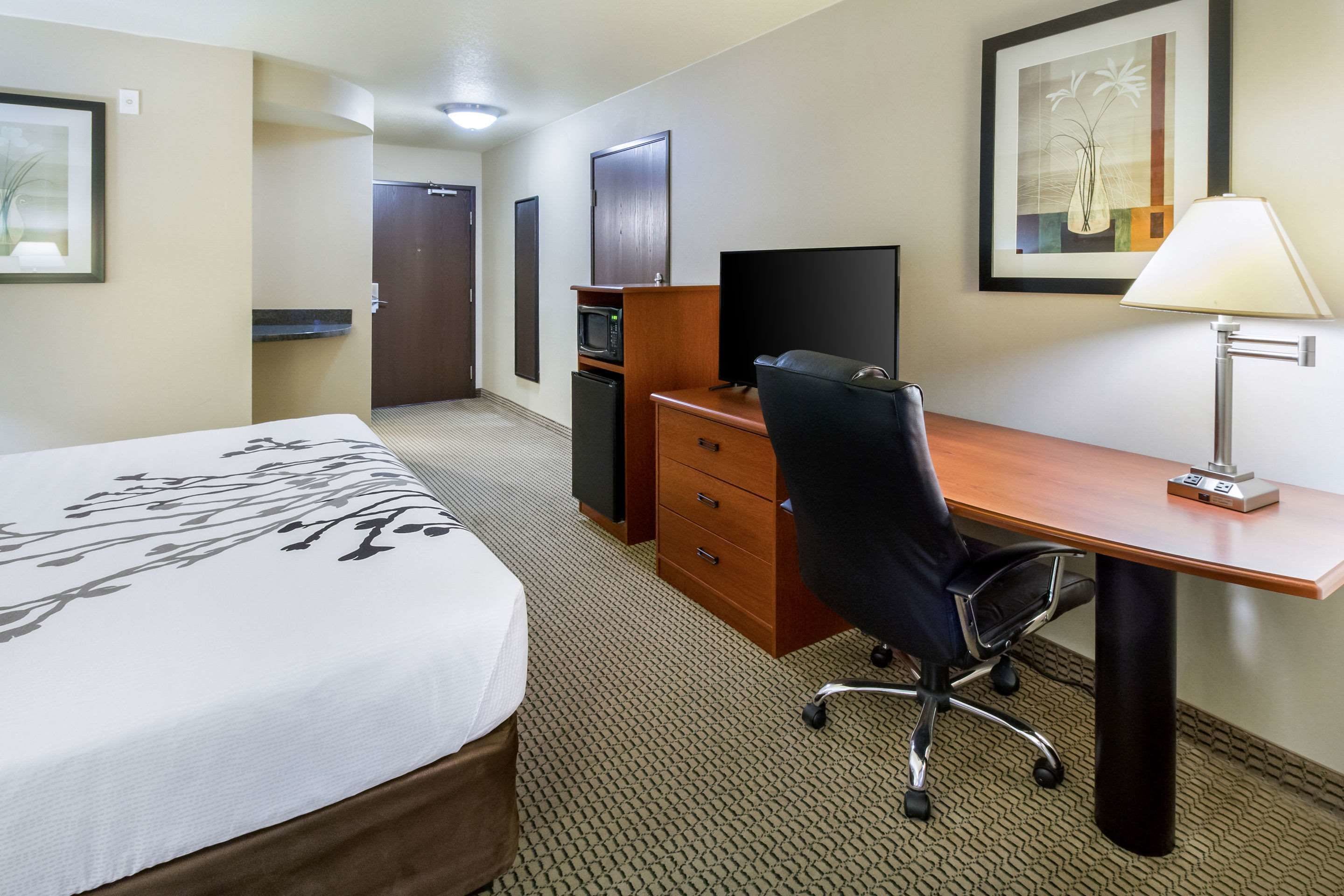 Una habitación de hotel con cama, escritorio con computadora, silla, lámpara, microondas y puerta.