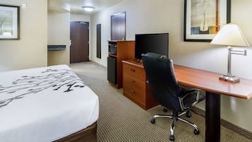 Una habitación de hotel con cama, escritorio con computadora, silla, lámpara, microondas y puerta.