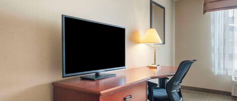A hotel room with a wooden desk, a flat-screen TV, a black office chair, a lamp, and a window with curtains.