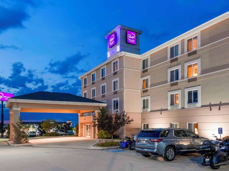 Un hotel con entrada cubierta, varios pisos y un estacionamiento con coches y motocicletas.