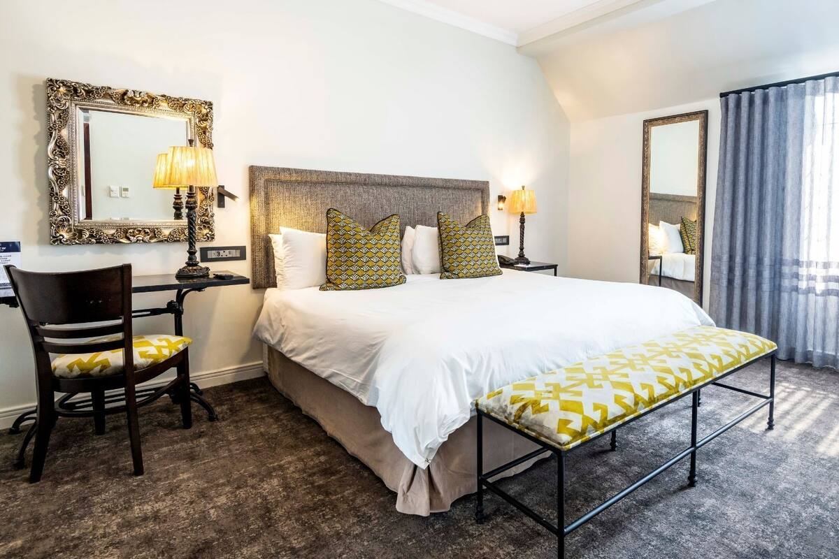 A hotel room with a large bed, two bedside tables with lamps, a chair, a bench, and two large mirrors on the walls.