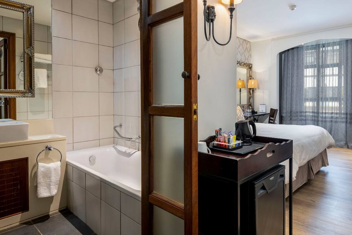 A hotel room with a bathtub, a desk with a coffee maker, a bed, and a large window with curtains.