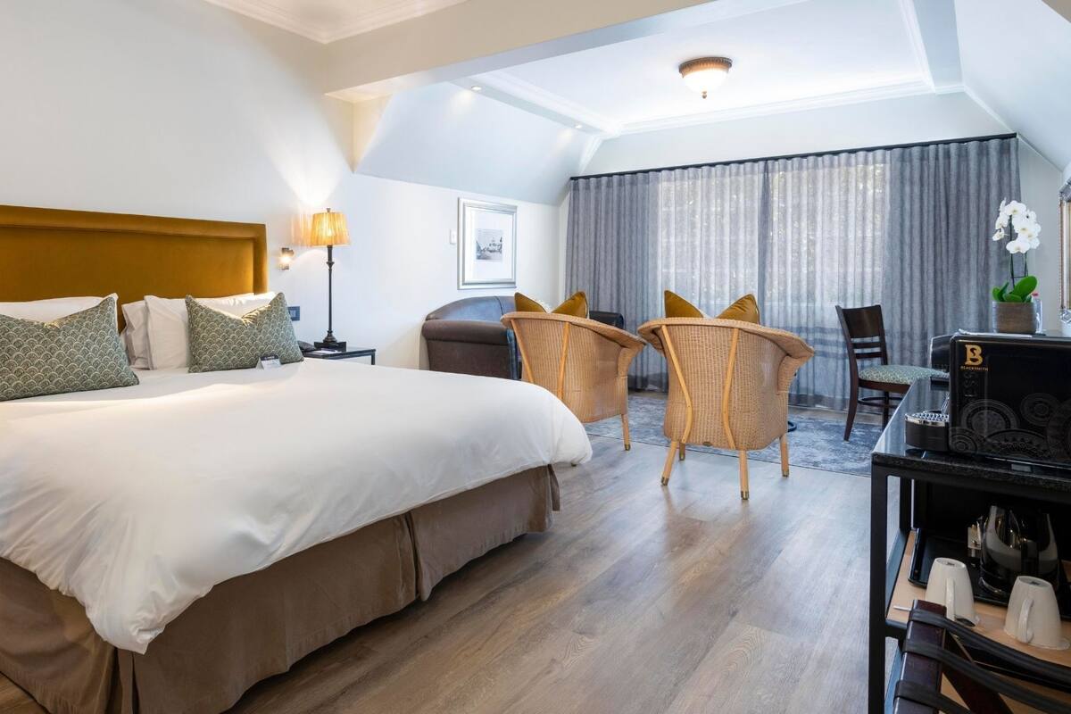 A hotel room with a large bed, two chairs, a desk with a coffee machine, and a window with curtains.