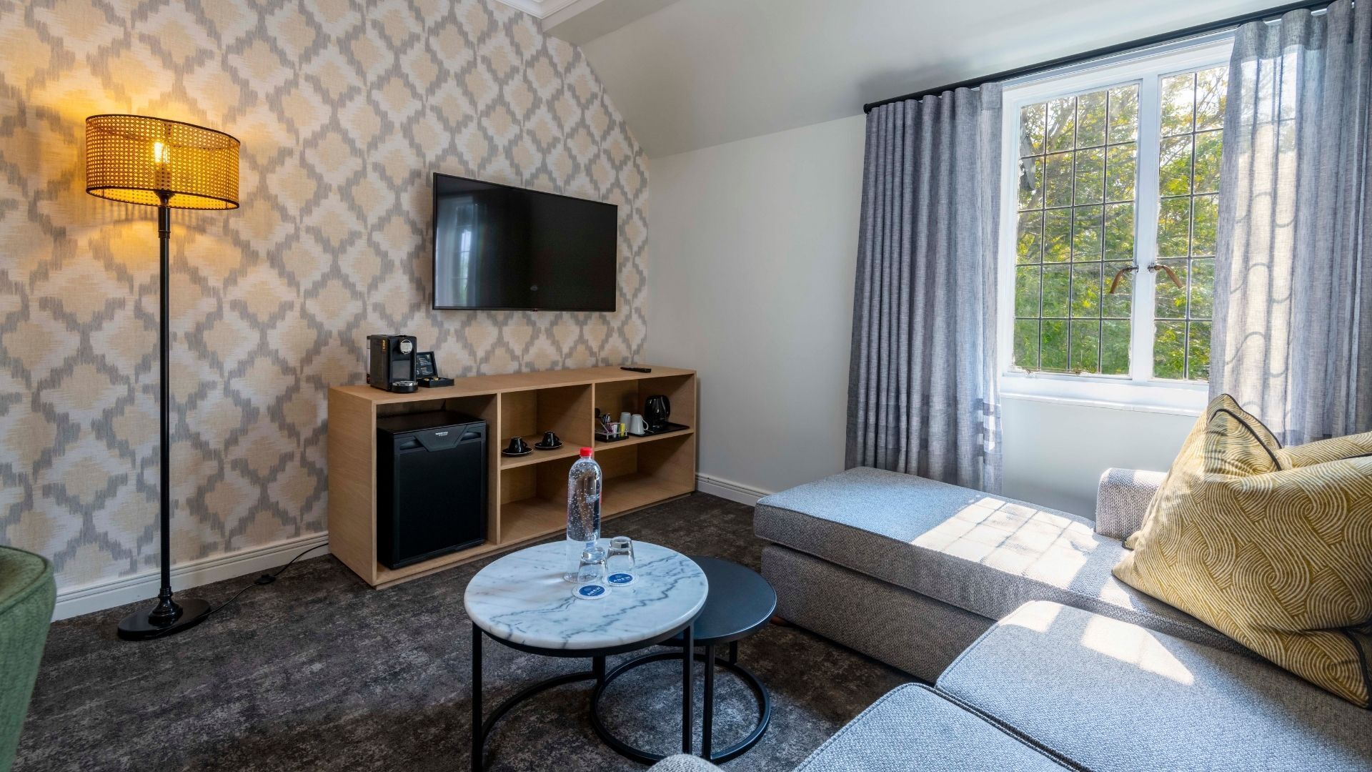 A modern living room with a flat-screen TV, a coffee machine, and a small table with a bottle and glass.