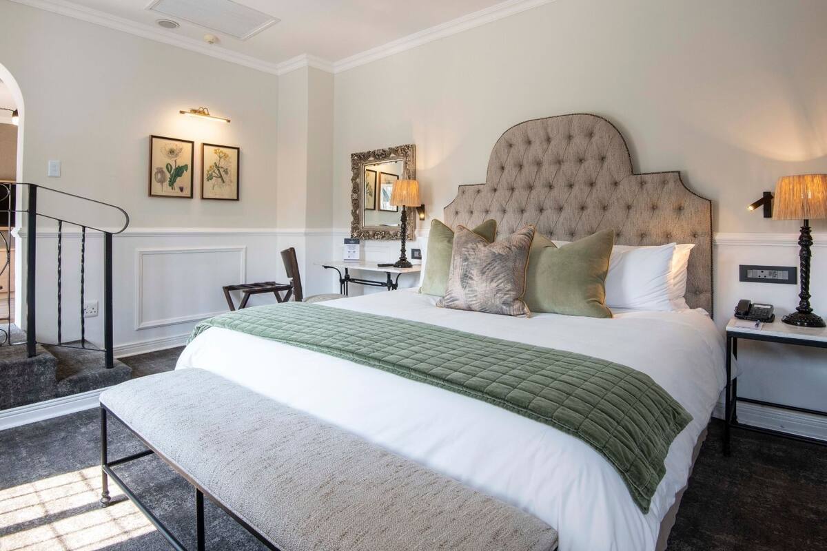 A hotel room with a large bed, a desk, a chair, a staircase, and a bathroom visible through an archway.