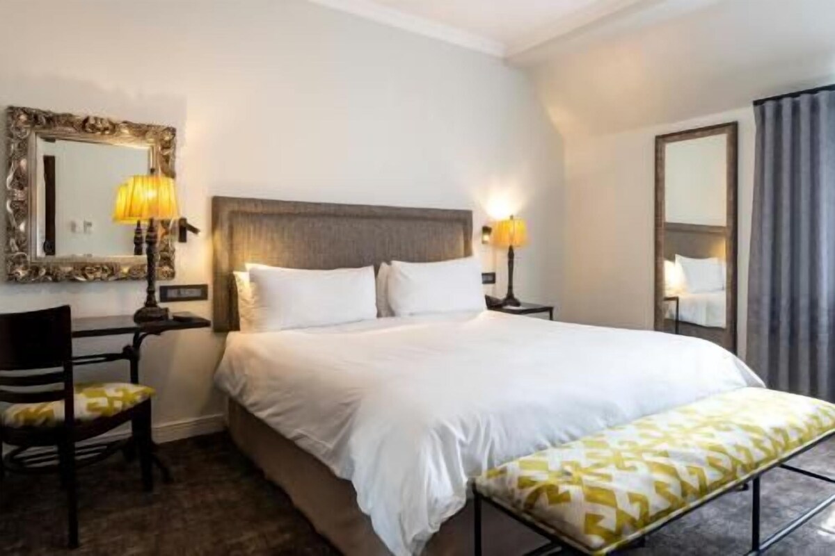 A hotel room with a large bed, two bedside tables with lamps, a chair, a bench, and a mirror on the wall.