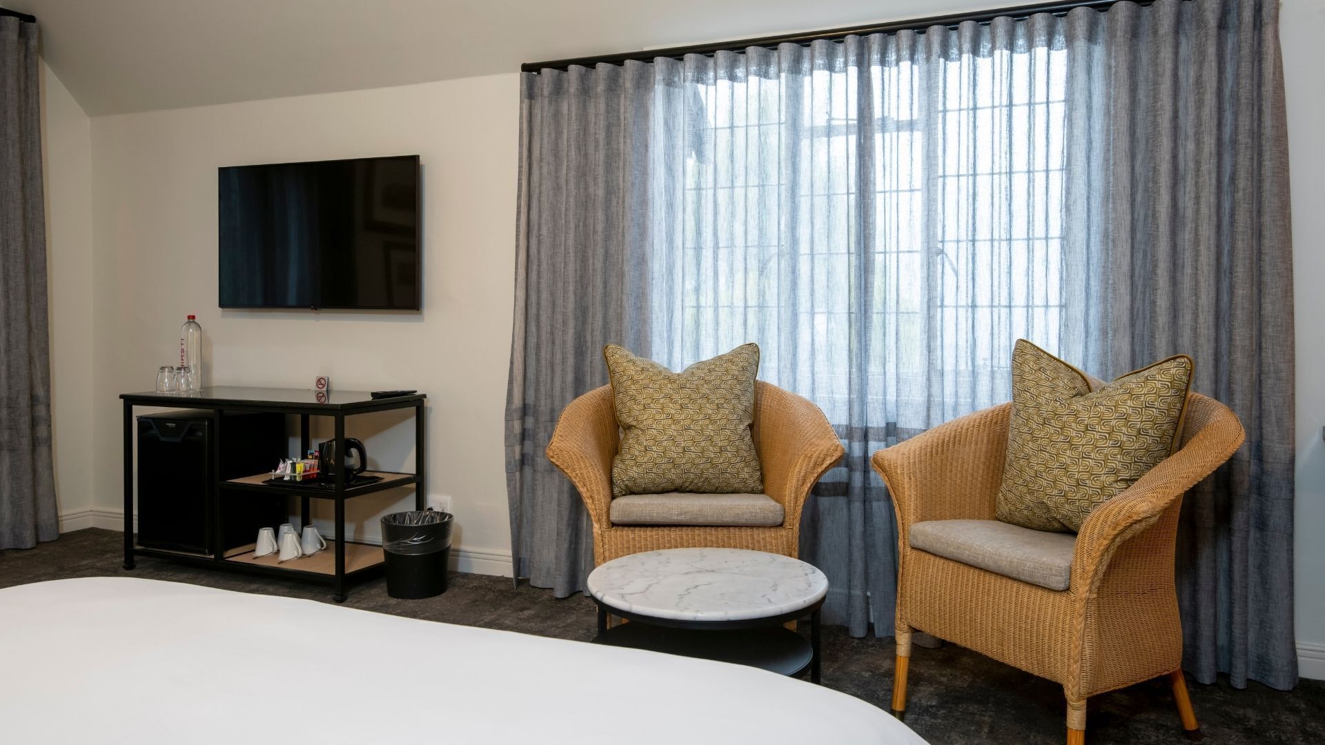 A hotel room with a bed, two wicker armchairs with patterned cushions, a small round table, a TV on the wall, and a minibar.