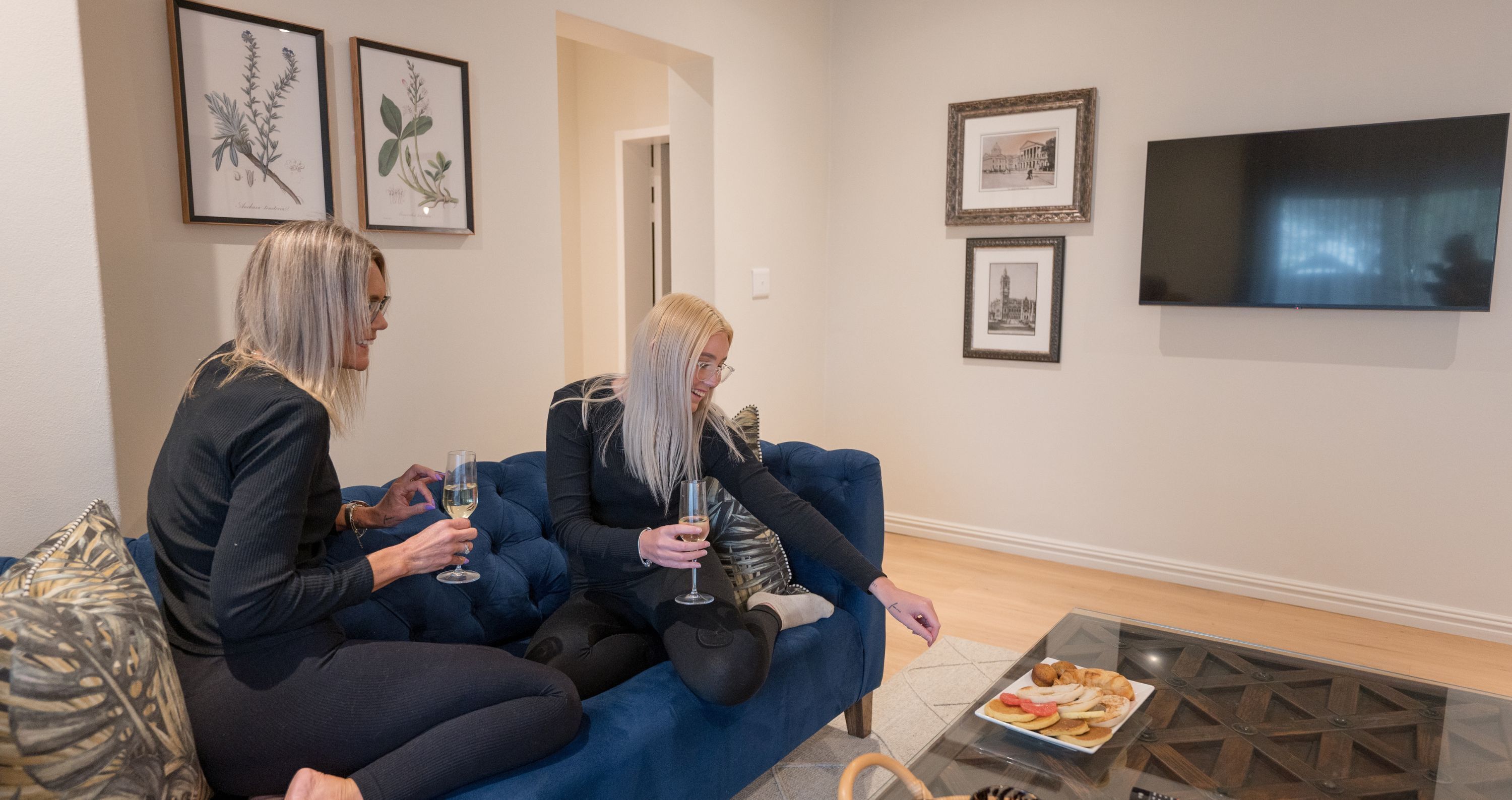Two people sitting on a blue sofa in a room with framed botanical prints on the wall, a flat-screen TV, and a coffee table with a plate of snacks.