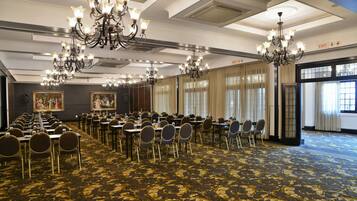 A conference room with multiple tables and chairs, chandeliers, and large windows with curtains.