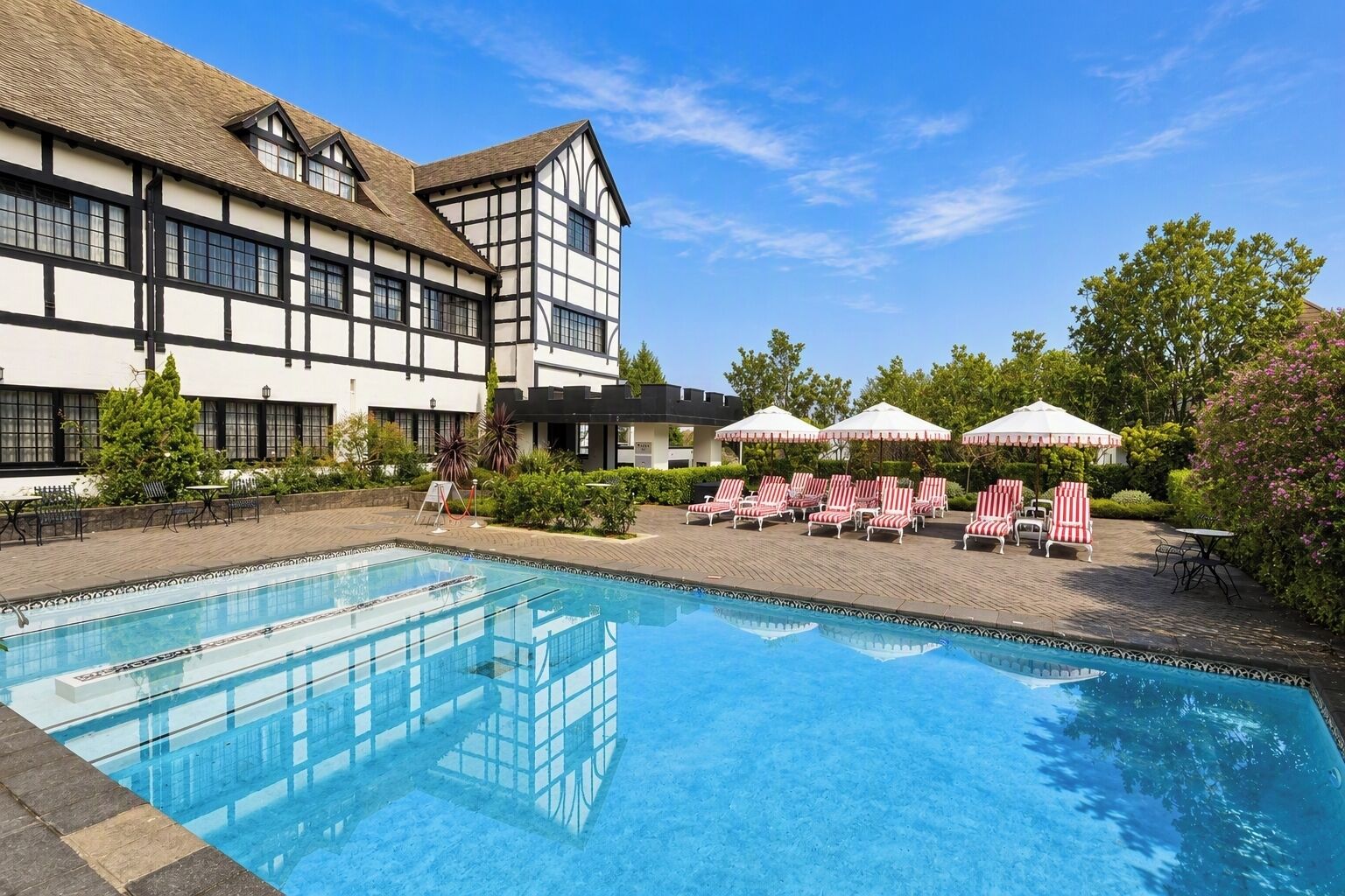 A pool area with lounge chairs and umbrellas, a building with a white facade and dark trim, and a clear blue sky.