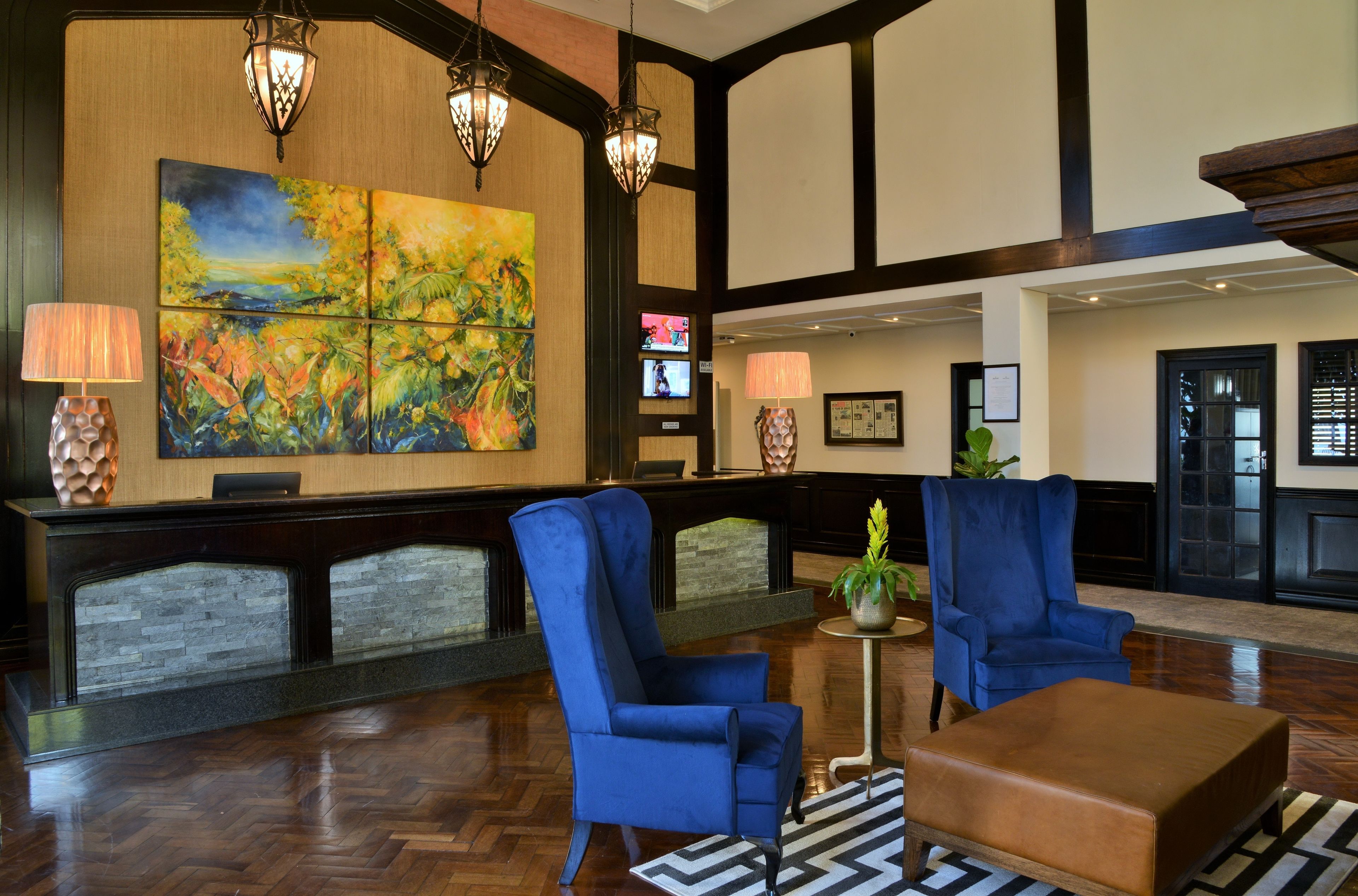 A lobby with two blue armchairs, a brown ottoman, a large abstract painting, and a reception desk with a lamp.