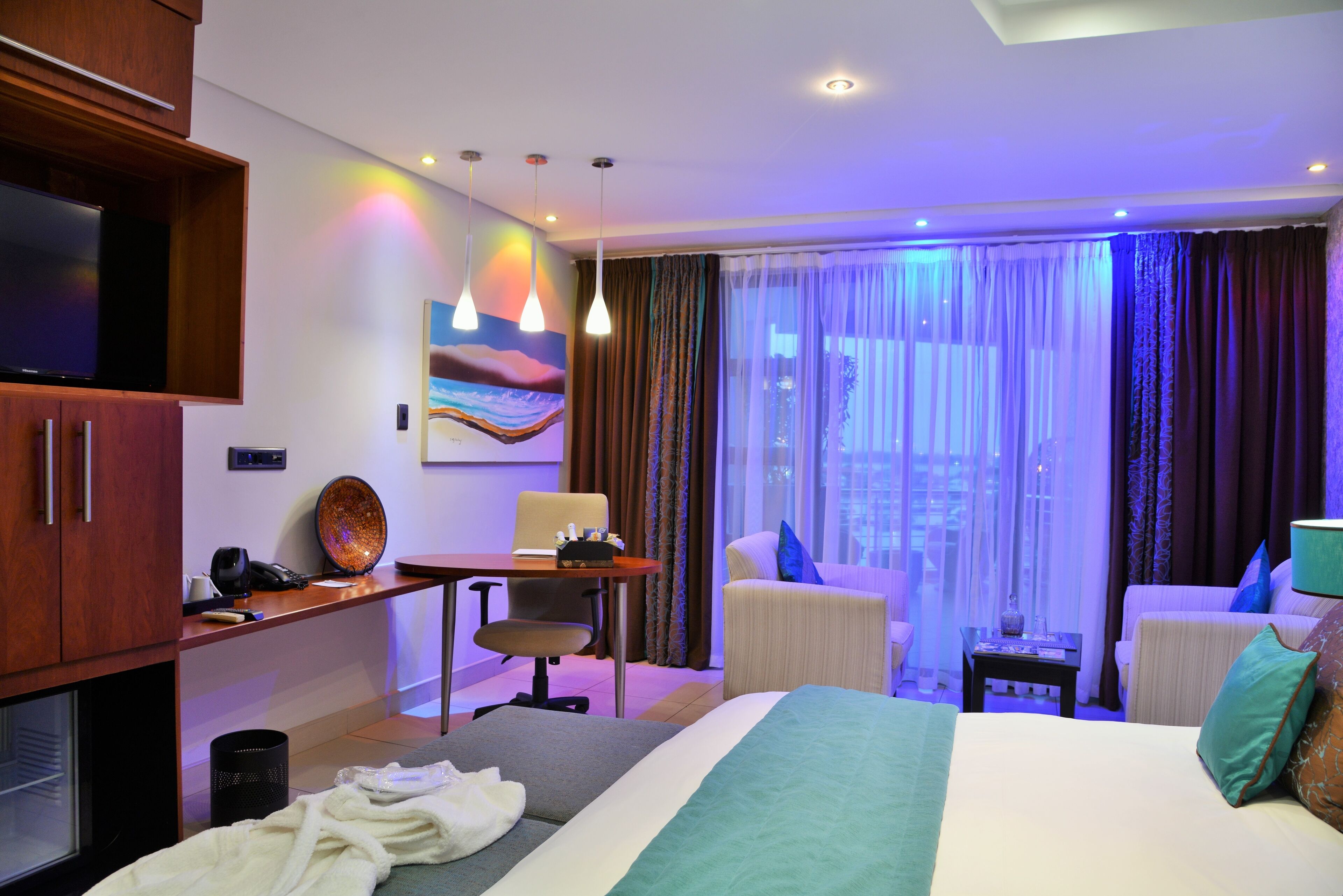 A hotel room with a large bed, a desk with a chair, a television, and a view of the city through the window.