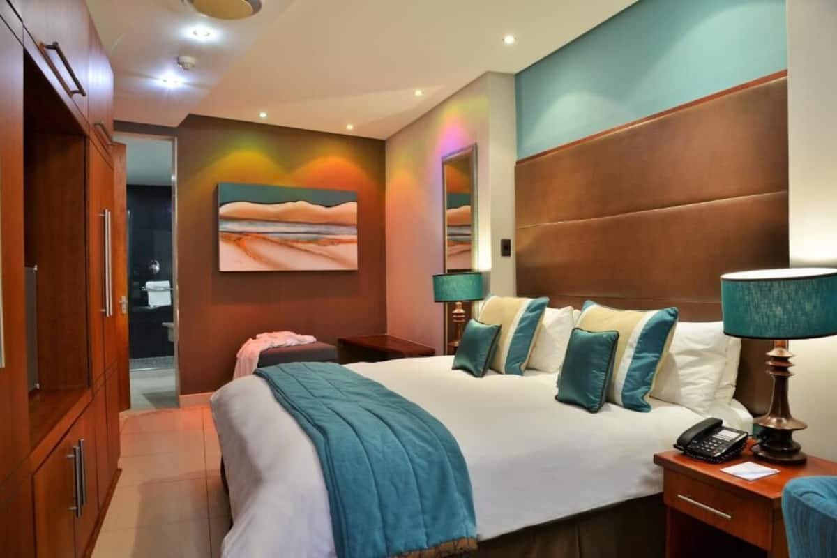 A hotel room with a large bed, a wooden wardrobe, a painting on the wall, and a bedside table with a lamp.