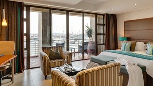 A hotel room with a balcony, a bed with multiple pillows, a coffee table, and striped armchairs. - BON Hotel Waterfront Richards Bay (Richards Bay)