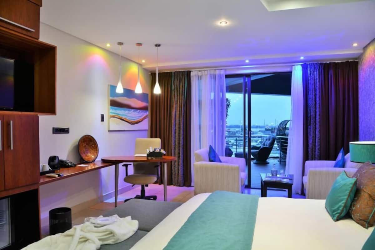 A hotel room with a bed, a desk, a chair, a TV, a painting, and a balcony with a view.
