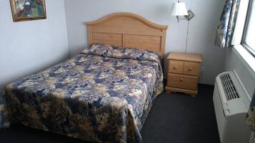 A bedroom with a wooden headboard, a bed with a floral bedspread, a bedside table with drawers, a wall-mounted lamp, and a window with curtains.