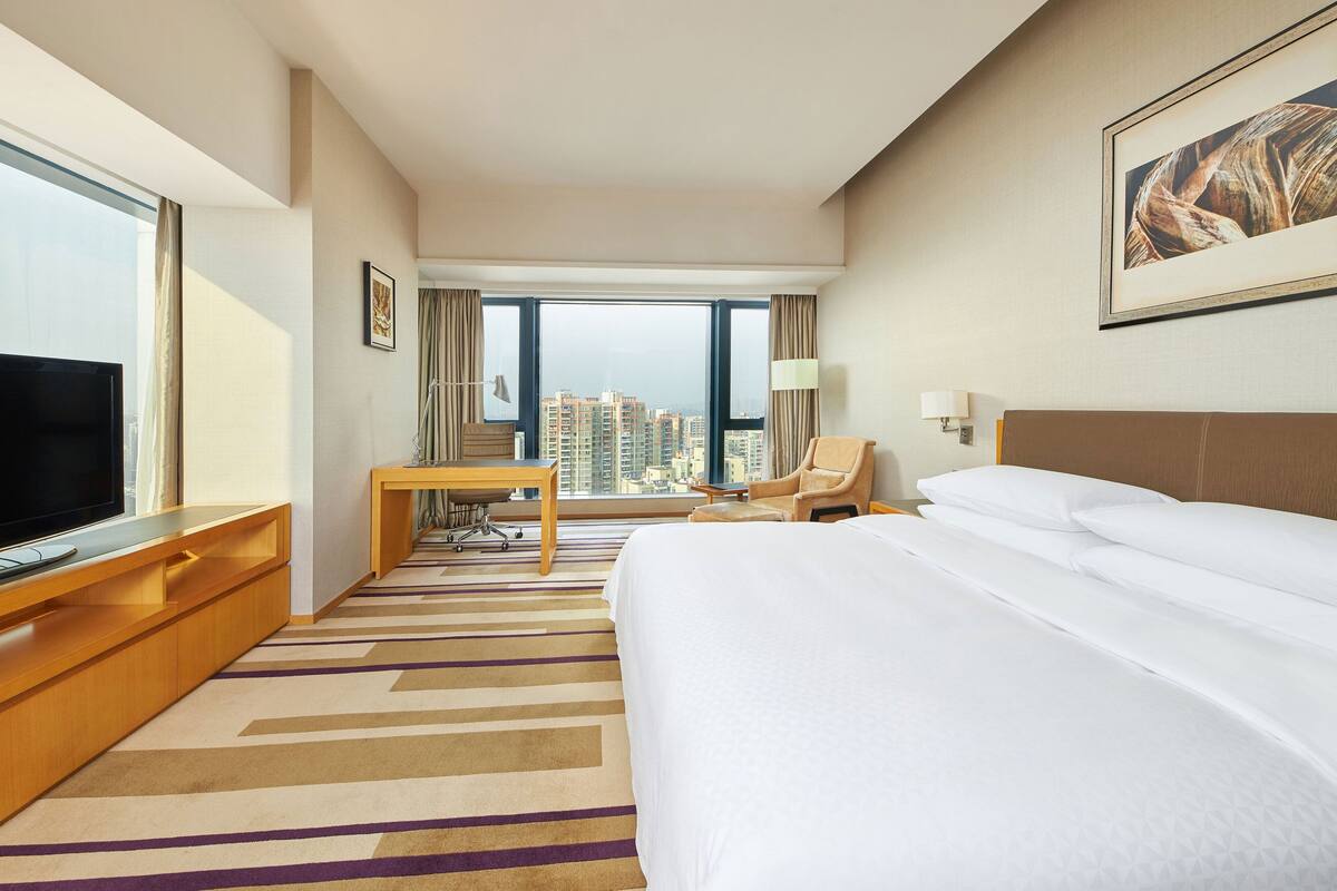 Quarto de hotel com uma cama grande, uma escrivaninha, uma cadeira, uma televisão e vista para a paisagem urbana.