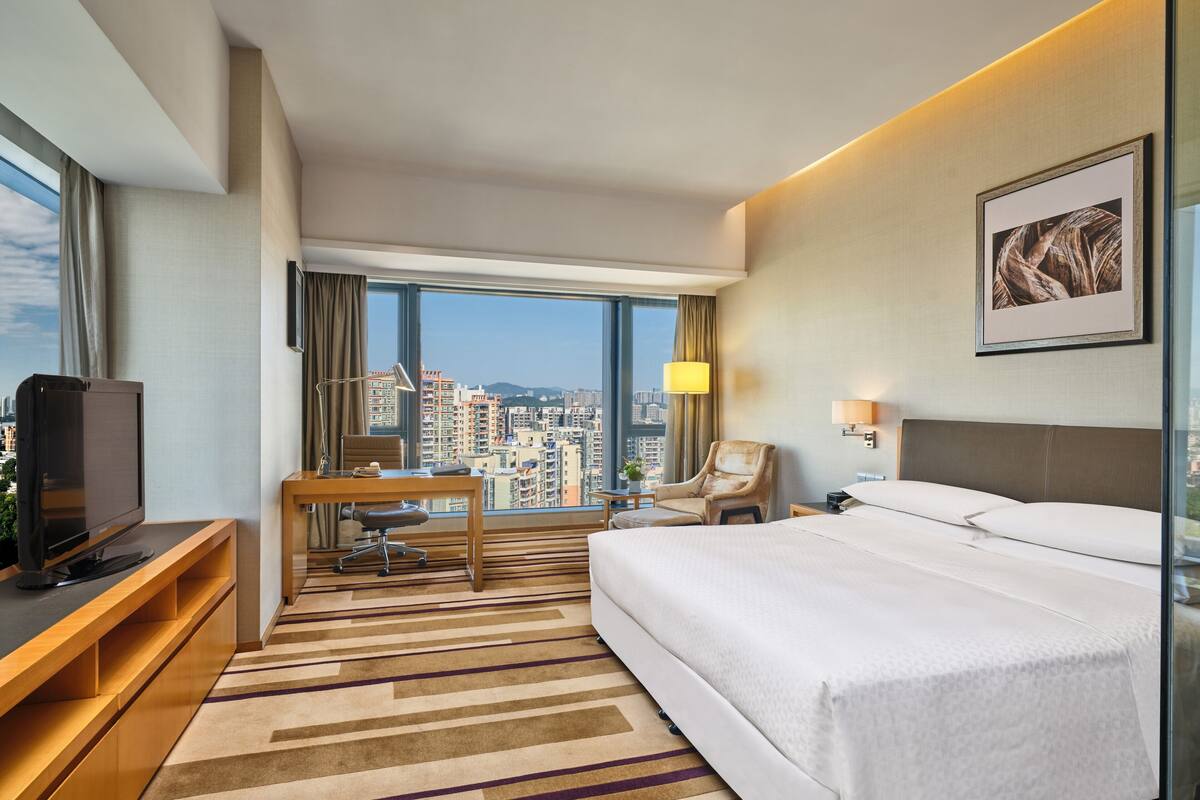 Quarto de hotel com cama, escrivaninha, cadeira, televisão e uma janela grande com vista para a cidade.