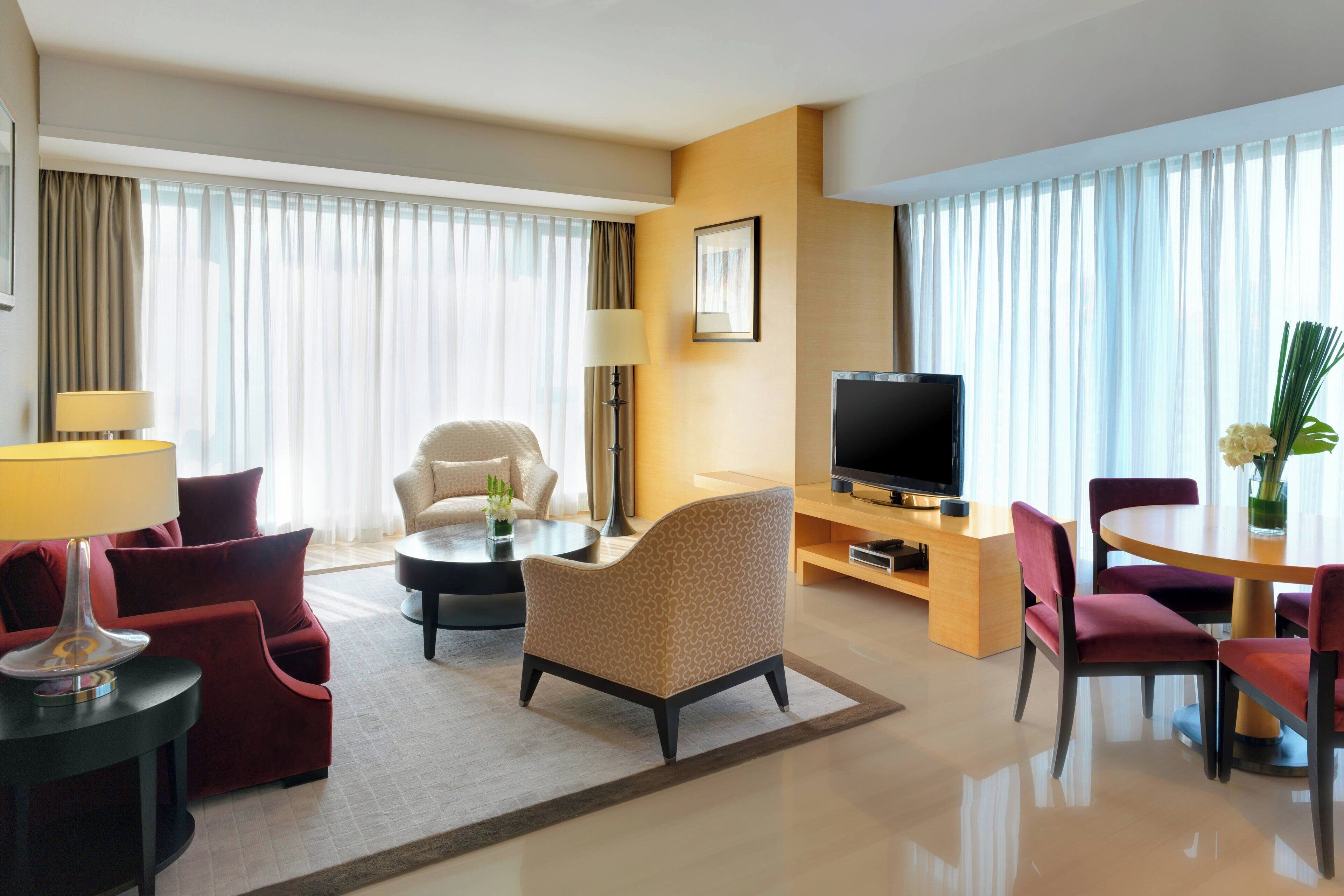 A hotel room with a sofa, armchairs, a TV, a dining table, and a lamp.