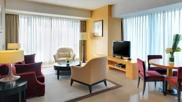 A hotel room with a sofa, armchairs, a TV, a dining table, and a lamp.