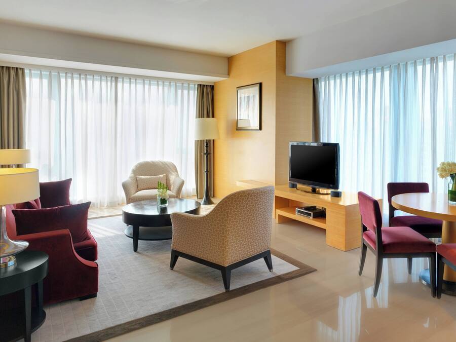 A hotel room with a sofa, armchairs, a TV, a dining table, and a lamp.