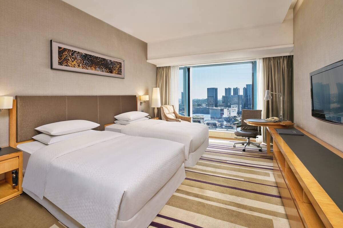 Quarto de hotel com duas camas, uma escrivaninha com cadeira, televisão e uma janela grande com vista para a cidade.