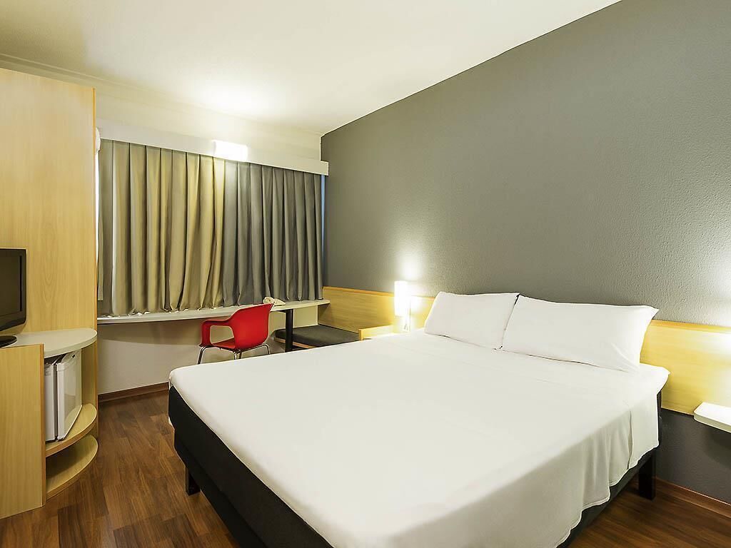 A hotel room with a large bed, a desk with a chair, a television, and a window with curtains.