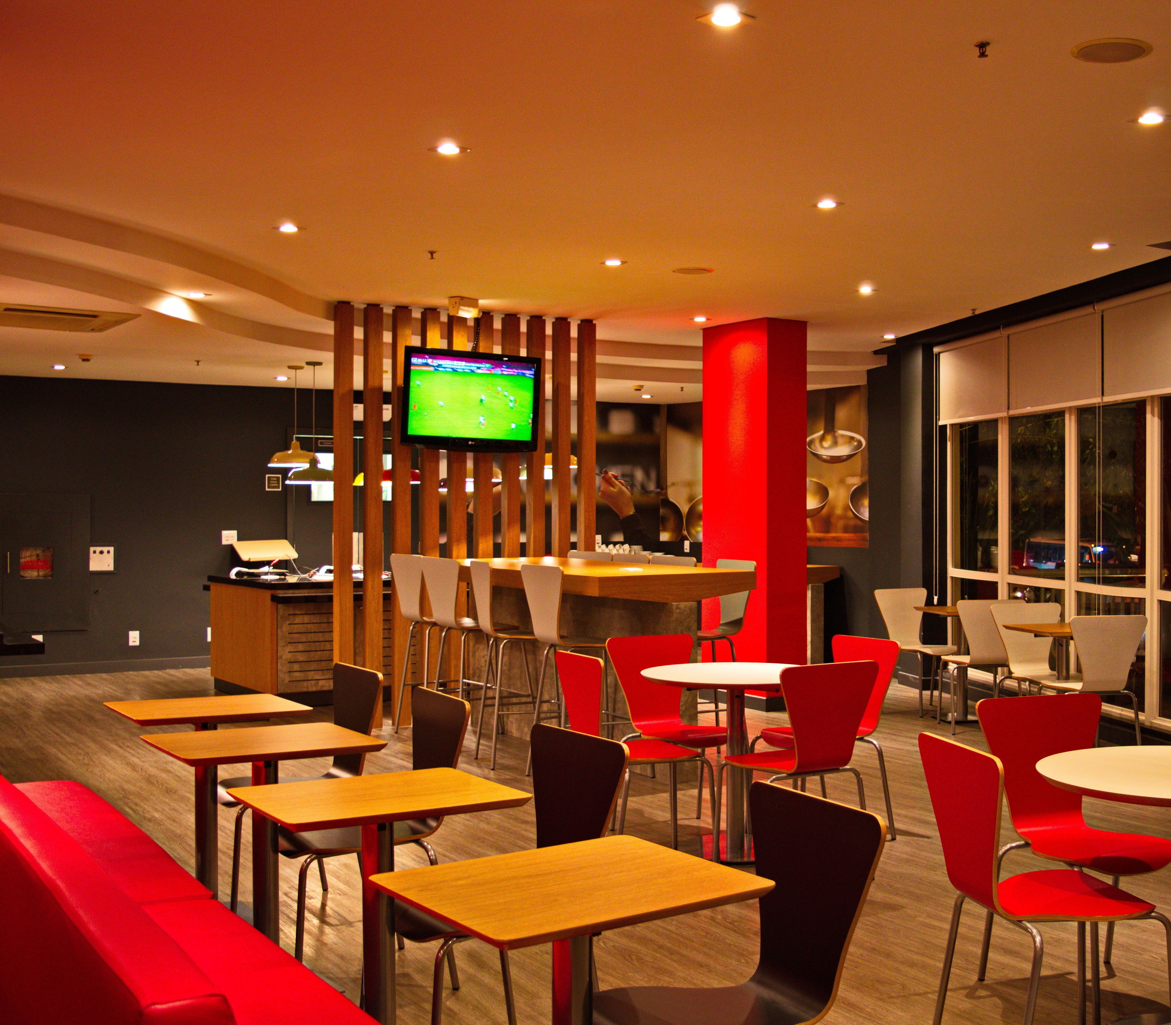 Un interior de restaurante moderno con varias mesas y sillas rojas, un televisor montado en una pared divisoria de madera y una zona de bar al fondo.