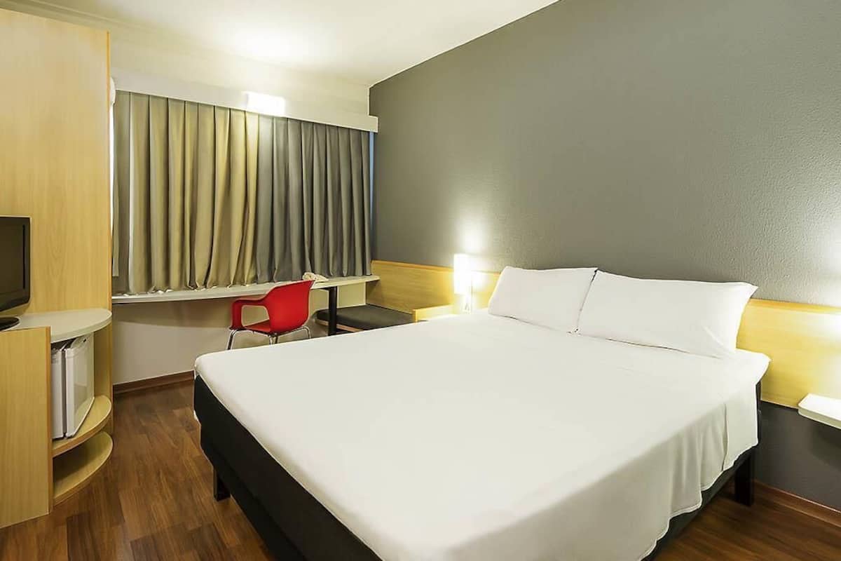 A hotel room with a large bed, a desk with a chair, a television, and a window with curtains.