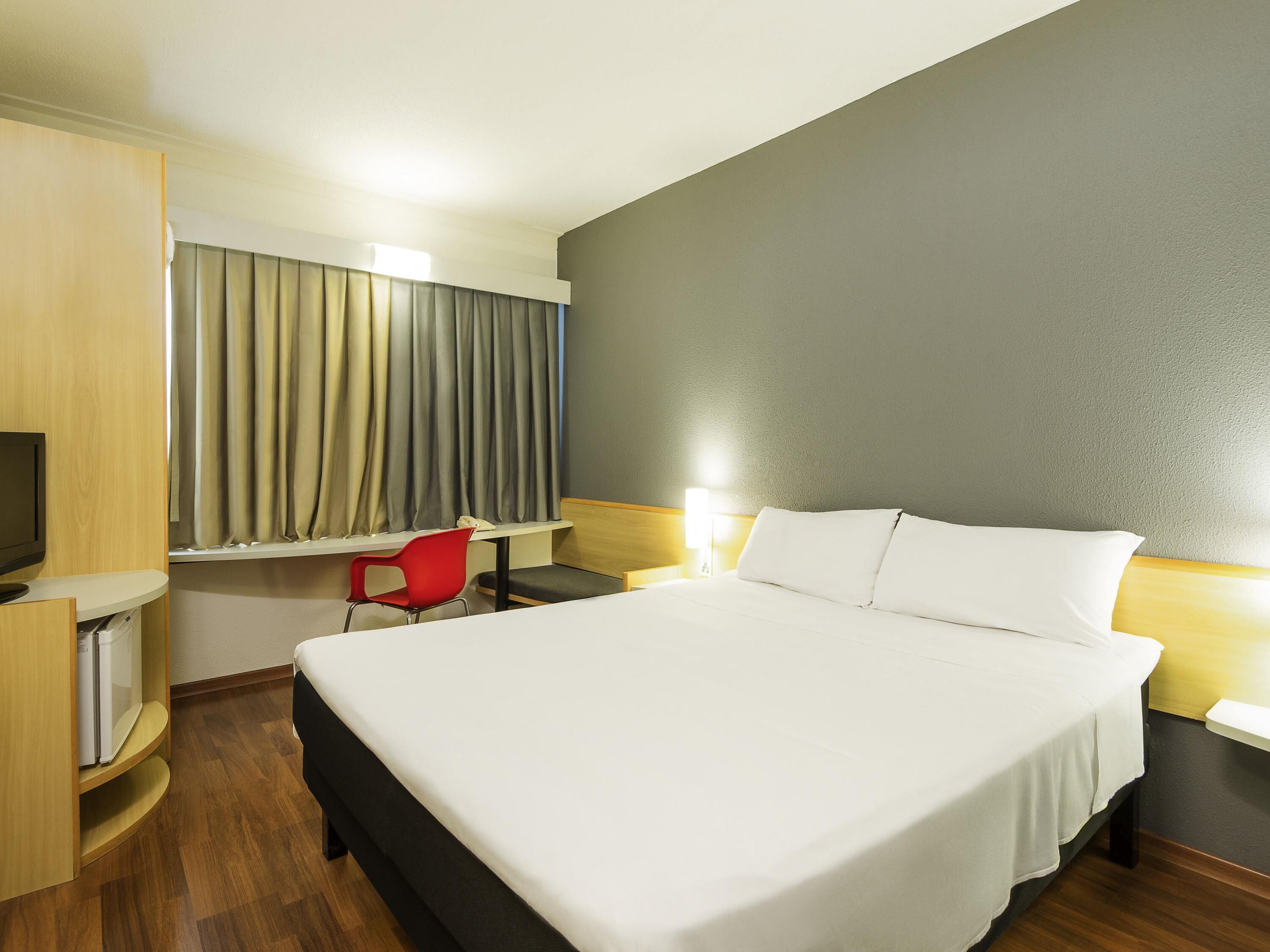 Une chambre d’hôtel avec un lit, un bureau, une chaise, une télévision et des rideaux.
