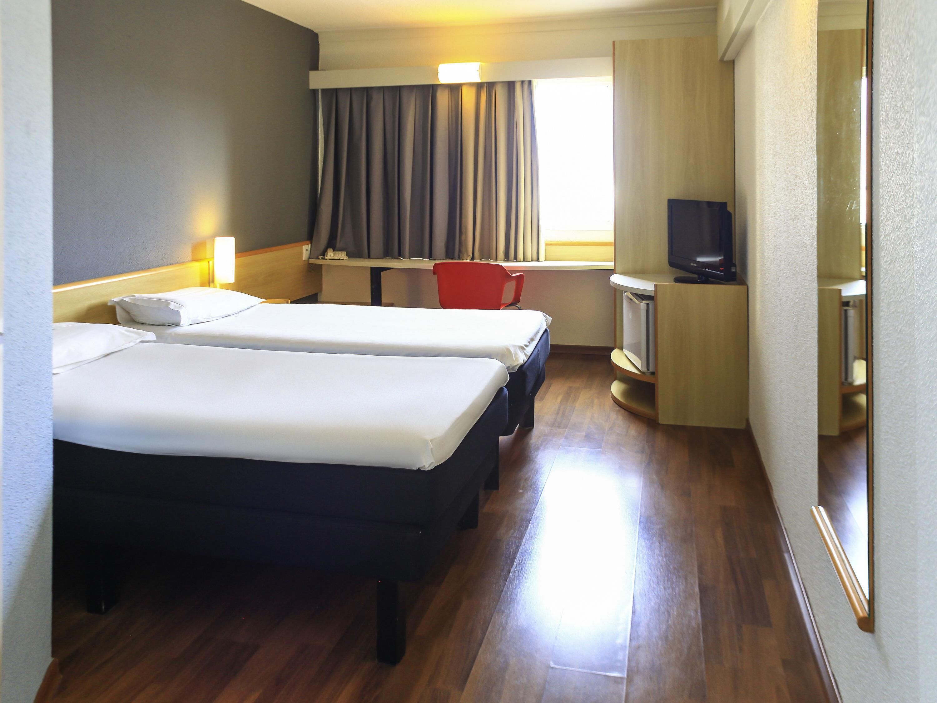 Habitación de hotel con dos camas, un escritorio con silla, televisor y ventana con cortinas.