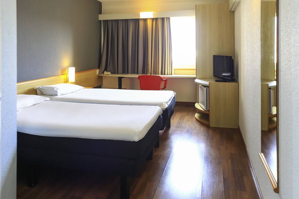 Habitación de hotel con dos camas, un escritorio con silla, televisor y ventana con cortinas.