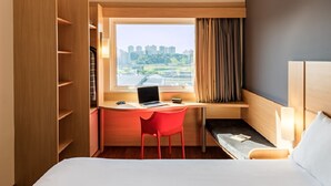 Habitación de hotel con cama, escritorio con computadora portátil, una silla roja y ventana con vista a edificios y vegetación.