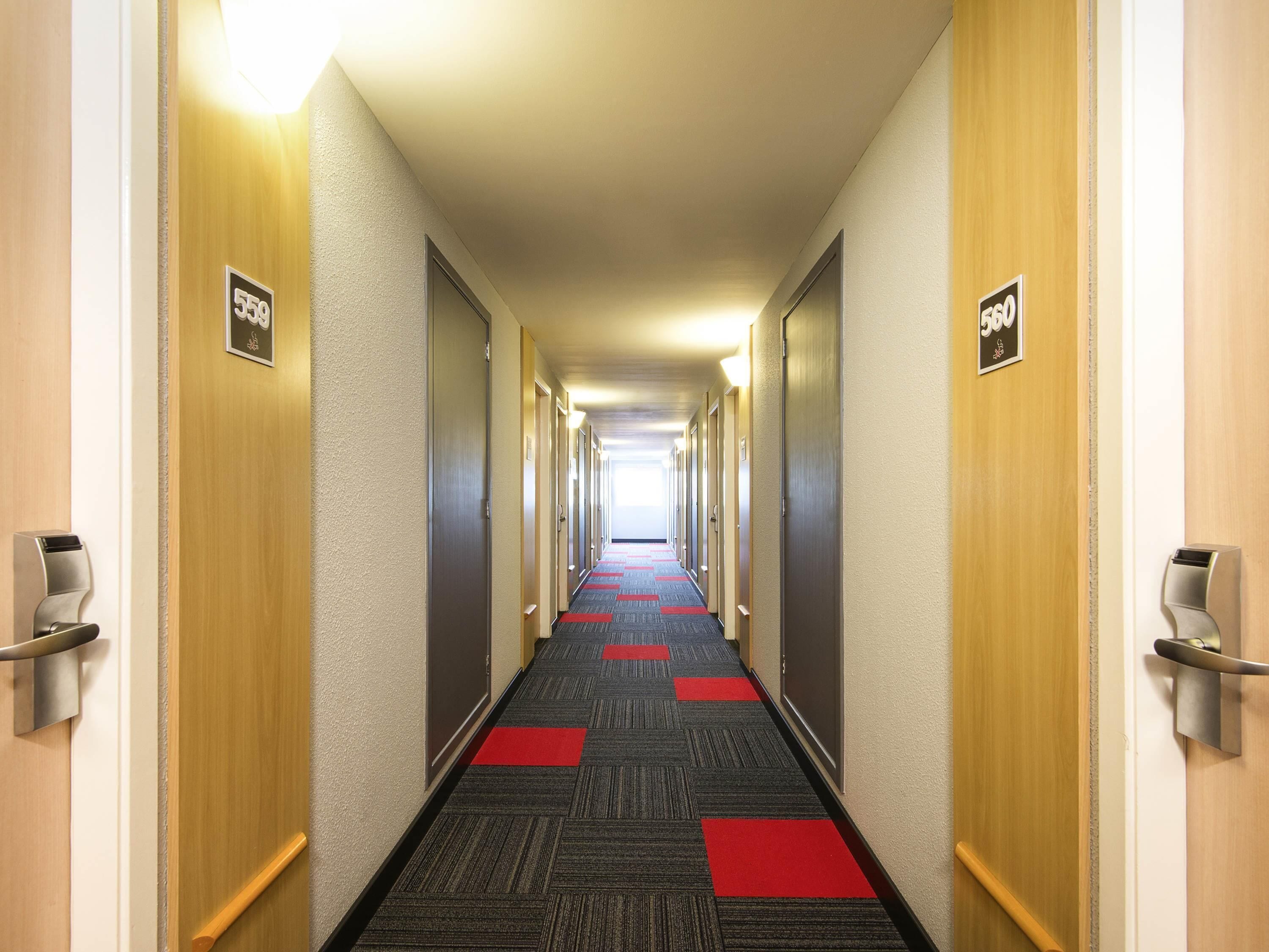 Un couloir d’hôtel avec des portes portant les numéros des chambres, un sol recouvert de moquette et des luminaires fixés aux murs.