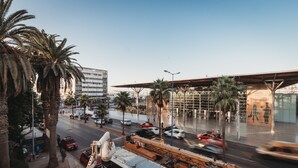 Una calle urbana concurrida con palmeras, un edificio moderno con fachada de vidrio y un sitio de construcción con una grúa y un camión que dice 'EXPRESS'.