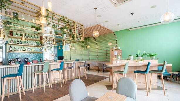 Una cafetería moderna con pared verde, barra de madera y varias zonas de estar.