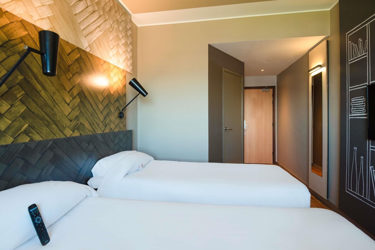 Ein Hotelzimmer mit zwei Betten, einer Wand mit Fischgrätmuster, einer Wandleuchte und einer Tür zu einem weiteren Raum.