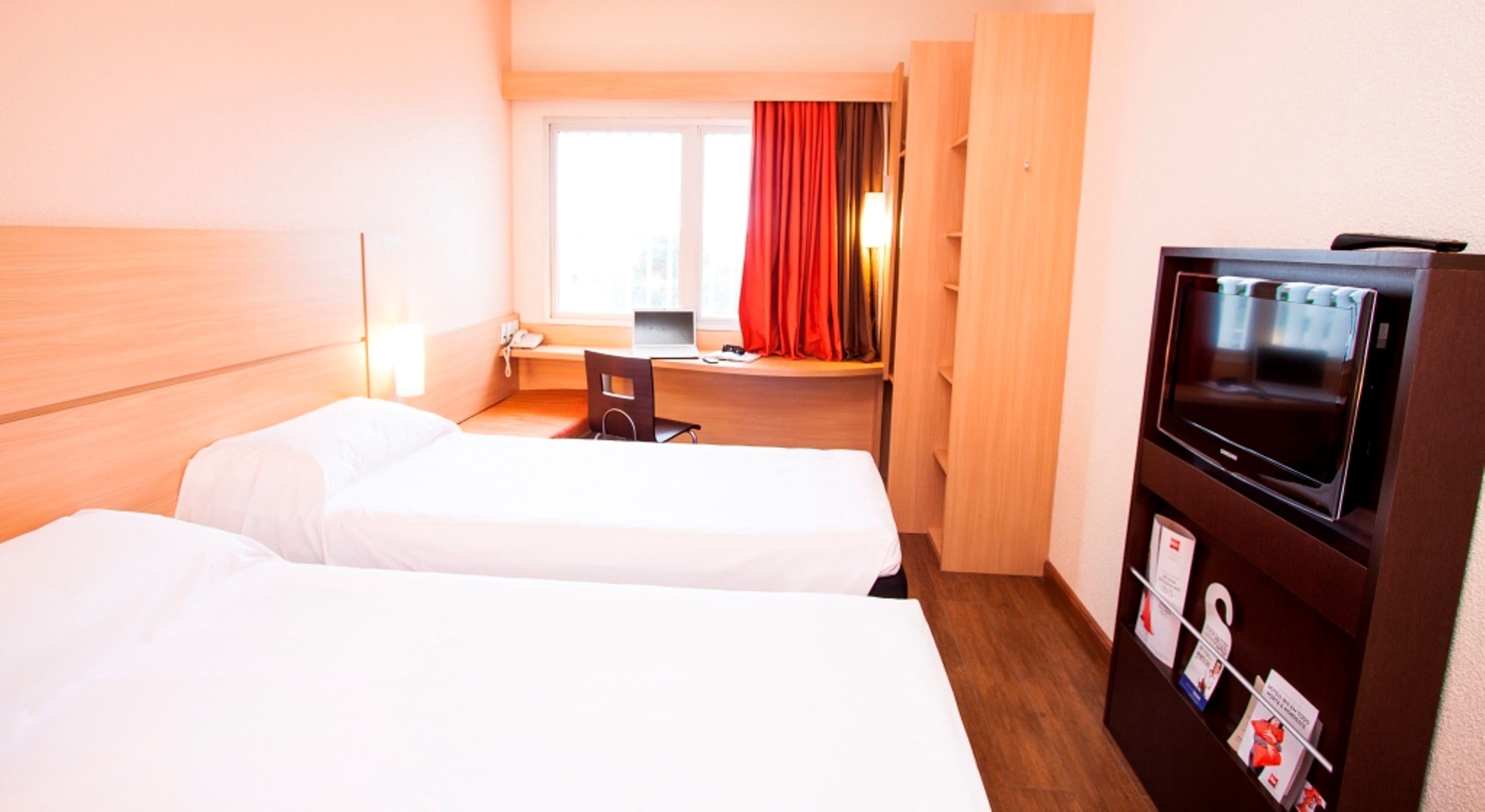 A hotel room with two beds, a desk, a TV, and a window with curtains.