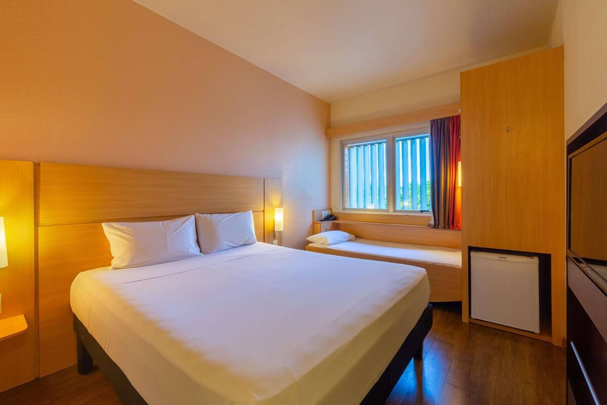 A hotel room with a large bed, a smaller bed, a desk, a chair, a window with curtains, and a mini-fridge.