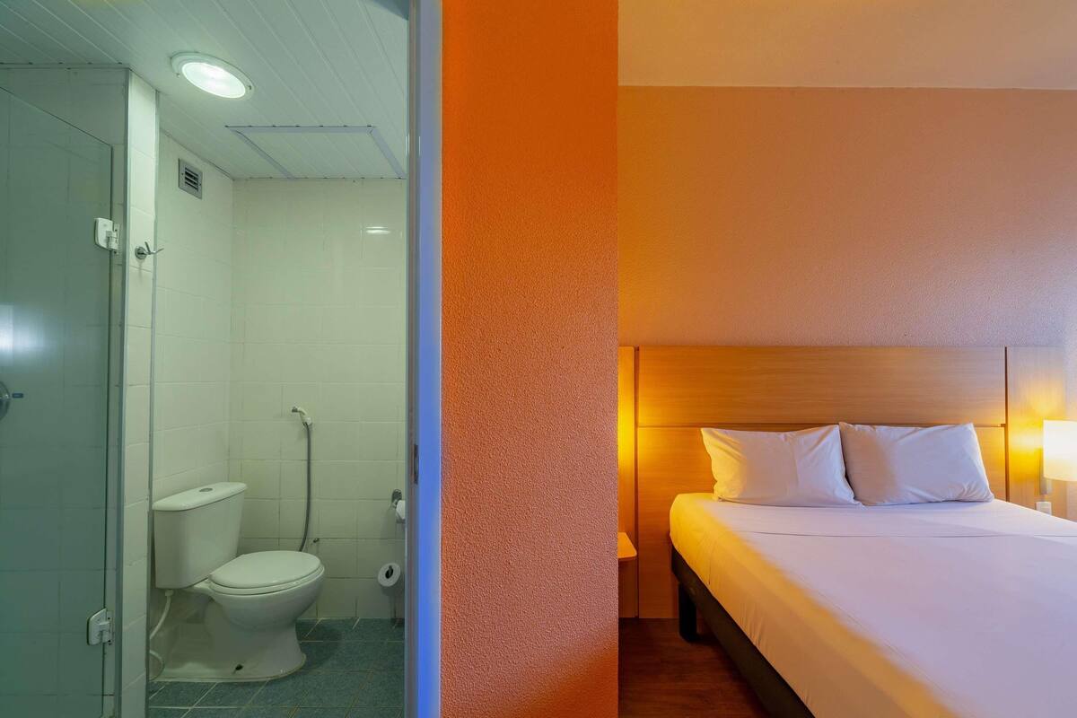 A hotel room with a shower, toilet, and a bed with a wooden headboard and two pillows.