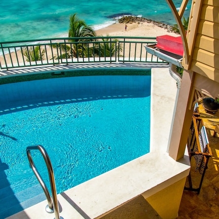 A swimming pool with a view of the ocean, a beach, and a patio area with chairs and a table.