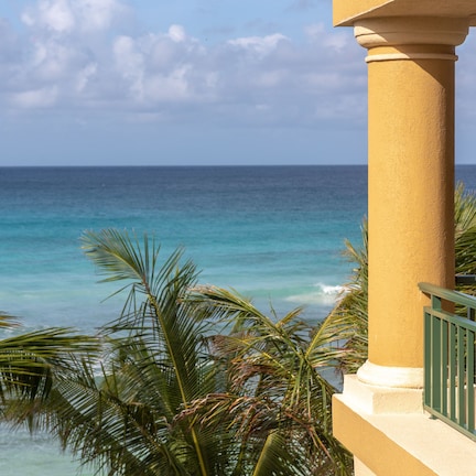 A balcony with a view of the ocean, palm trees, and a building with a beige facade and green railing.