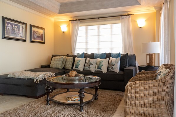 A living room with a sofa, a round coffee table, and a wicker chair. There are two framed pictures on the wall and a window with curtains.