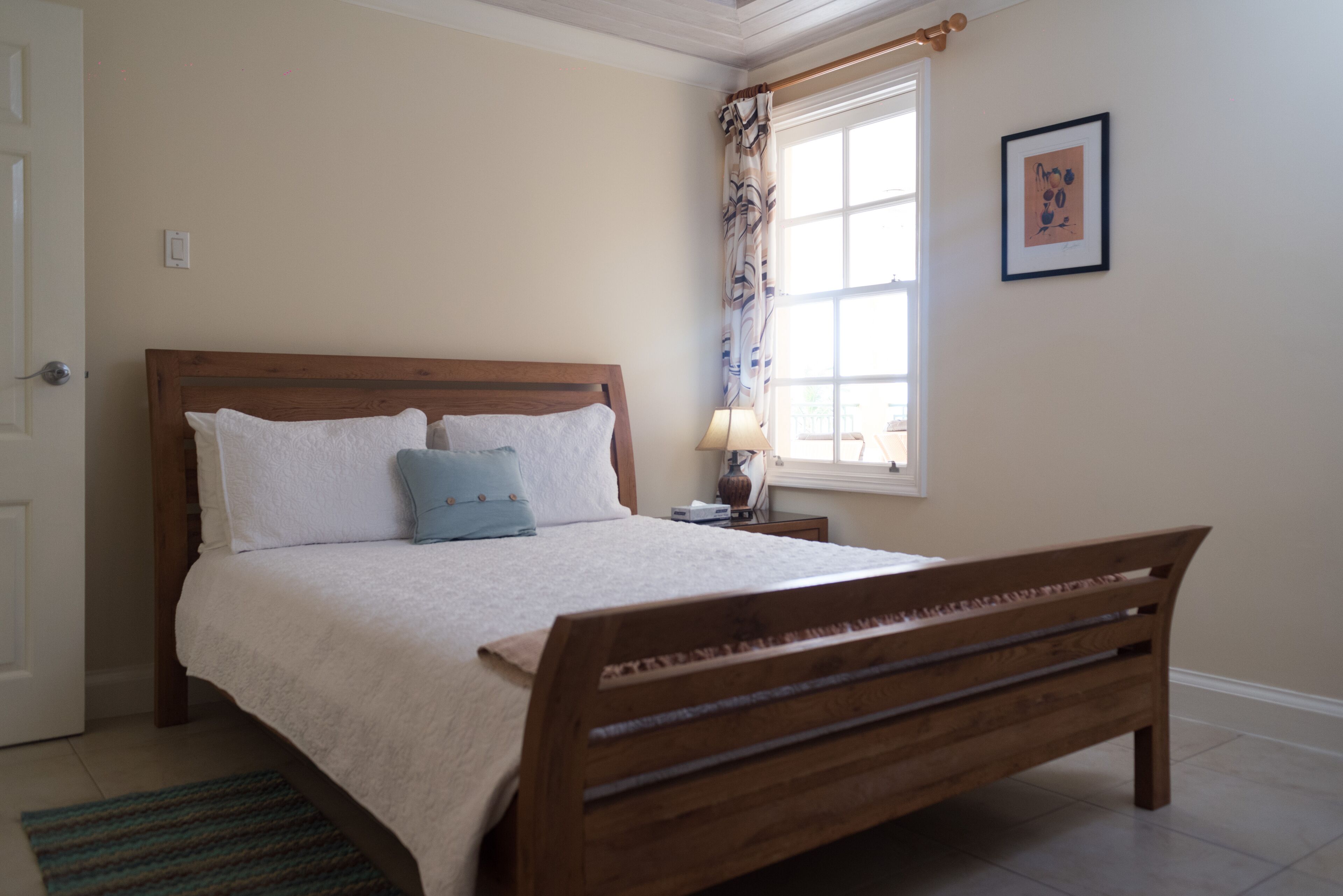 A bedroom with a wooden bed, a nightstand, a lamp, a window with curtains, and a framed picture on the wall.