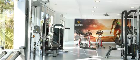 Un gimnasio moderno con diversas máquinas de ejercicio, un gran cartel motivacional y vistas a palmeras afuera.