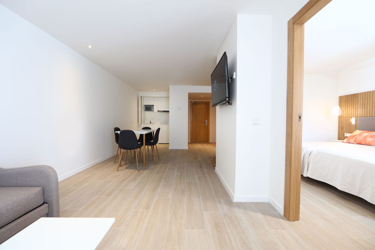 Ein modernes Hotelzimmer mit einem Bett, einem an der Wand befestigten Fernseher, einer Essecke mit Tisch und Stühlen sowie einer Küche im Hintergrund.