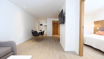 Una habitación de hotel moderna con una cama, un televisor montado en la pared, una zona de comedor con mesa y sillas, y una cocina al fondo.