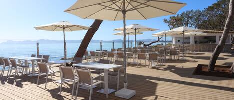 An outdoor deck with tables and chairs, large umbrellas, and a view of the sea.